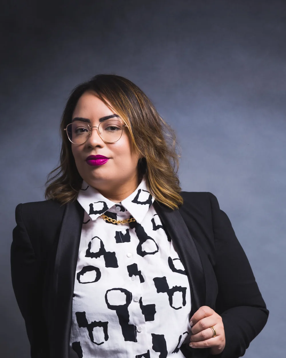 Amanda Noronha in a black blazer over a patterned blouse, studio portrait with a dark background