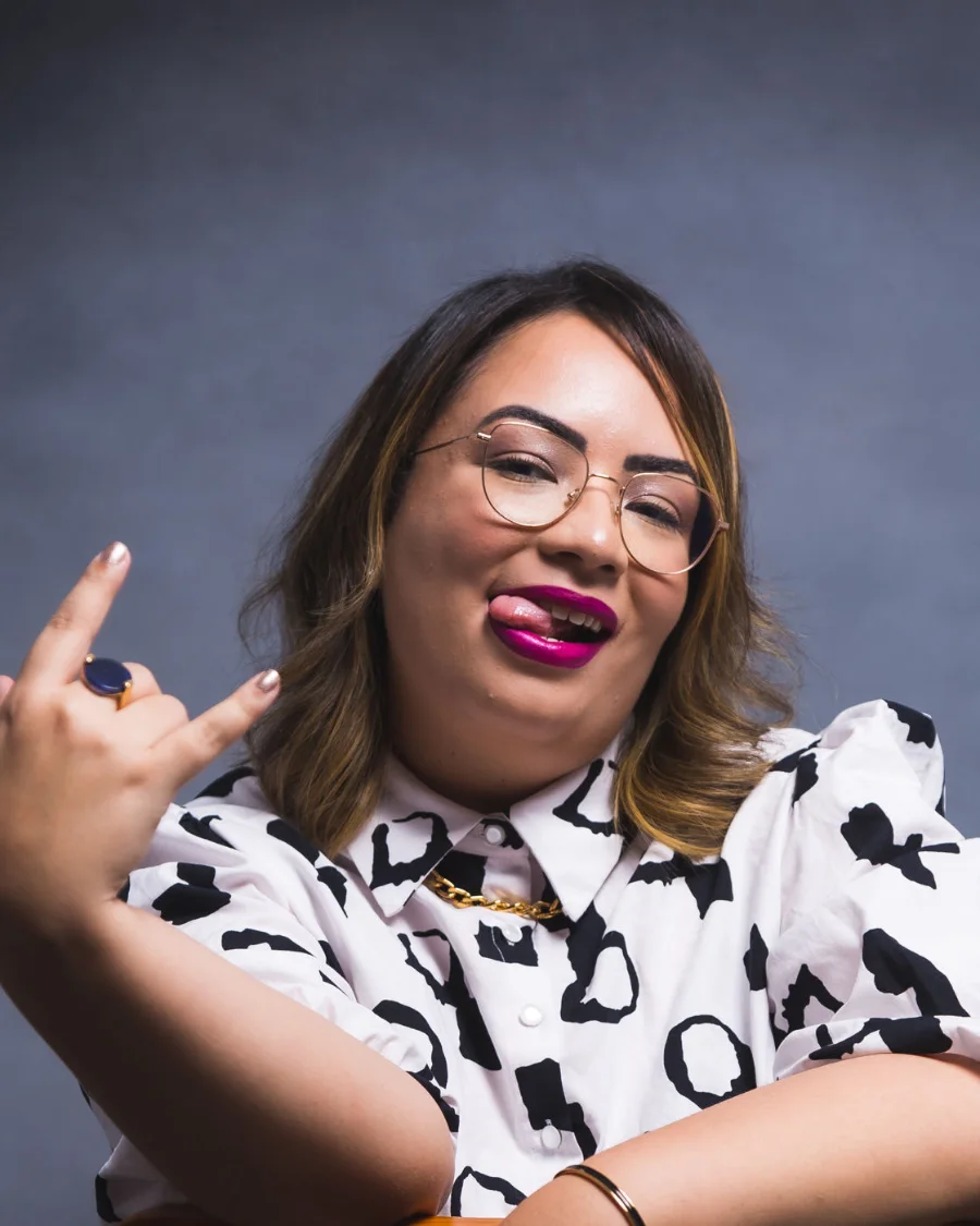 Amanda Noronha sitting backwards on a chair, smiling and making the rock sign with her hand
