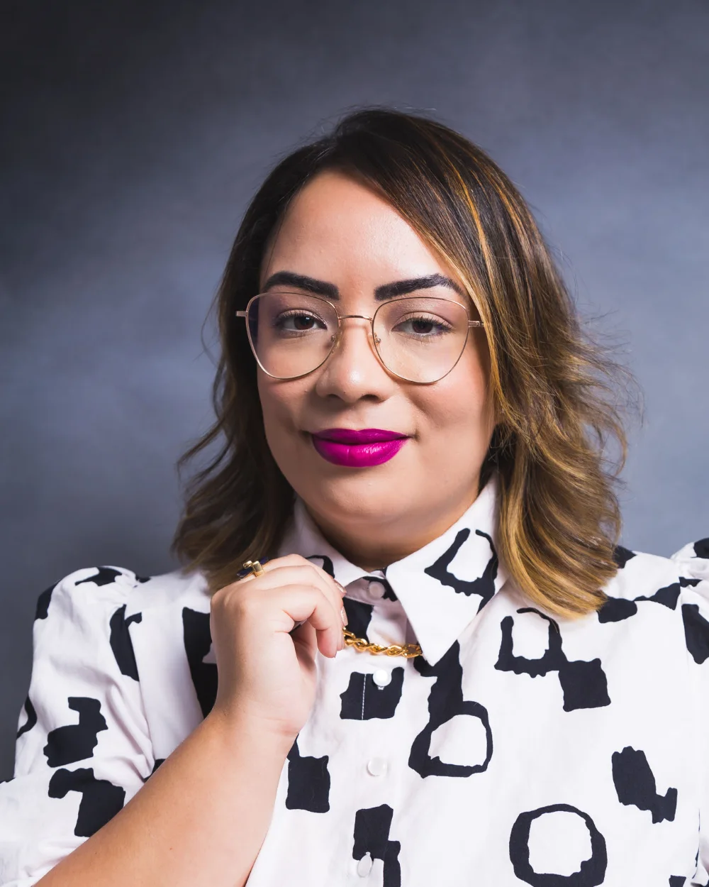 Amanda Noronha in a studio portrait, wearing glasses, magenta lipstick and a black and white patterned blouse
